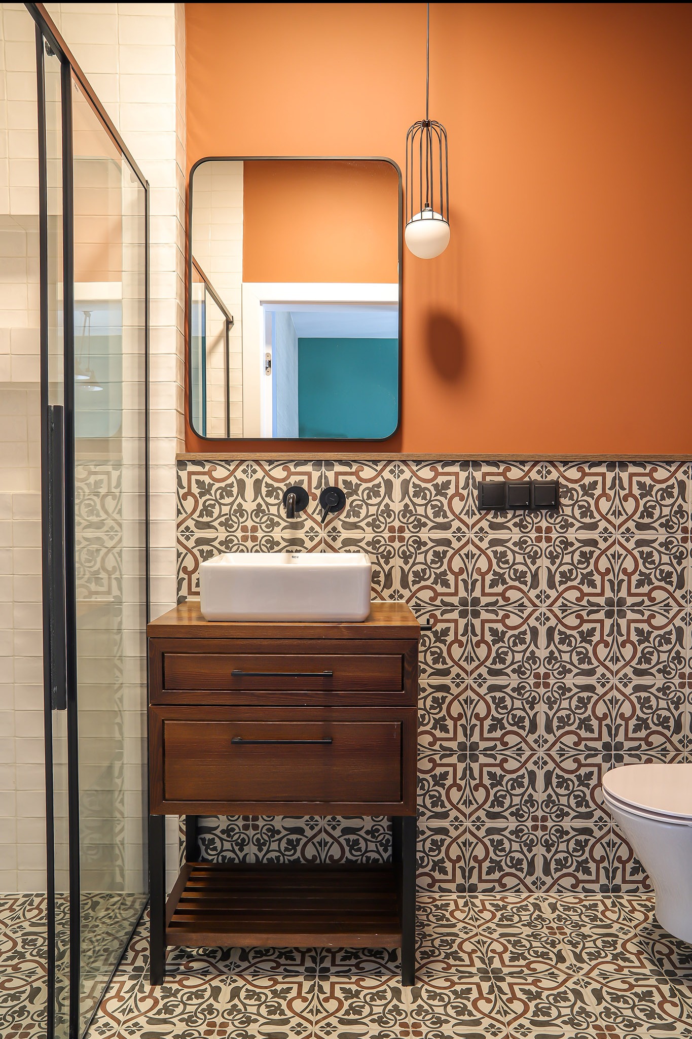 Nowoczesna łazienka z kabiną prysznicową, umywalką na szafce z drewna i wzorzystymi płytkami. Pomarańczowa ściana i designerska lampa wisząca.
