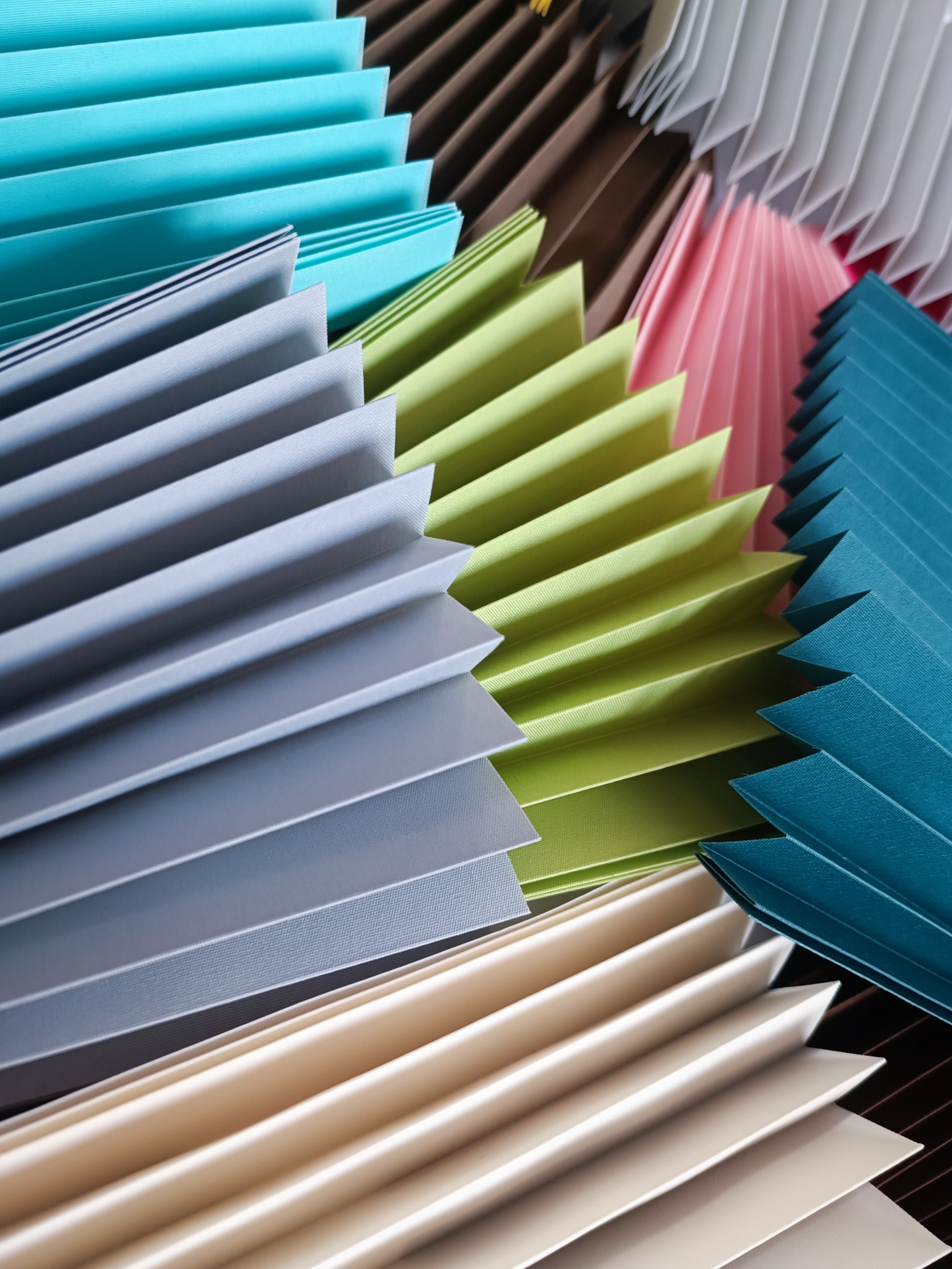 Kolorowe fragmenty plis okiennych w zbliżeniu: turkusowe, szare, zielone, różowe i beżowe, tworzące geometryczny wzór z wyraźną teksturą materiału.