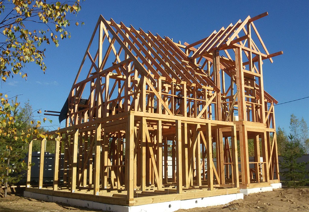 Dwukondygnacyjny dom szkieletowy w trakcie budowy, z widoczną drewnianą konstrukcją nośną i fragmentem fundamentów na tle błękitnego nieba i zieleni drzew.