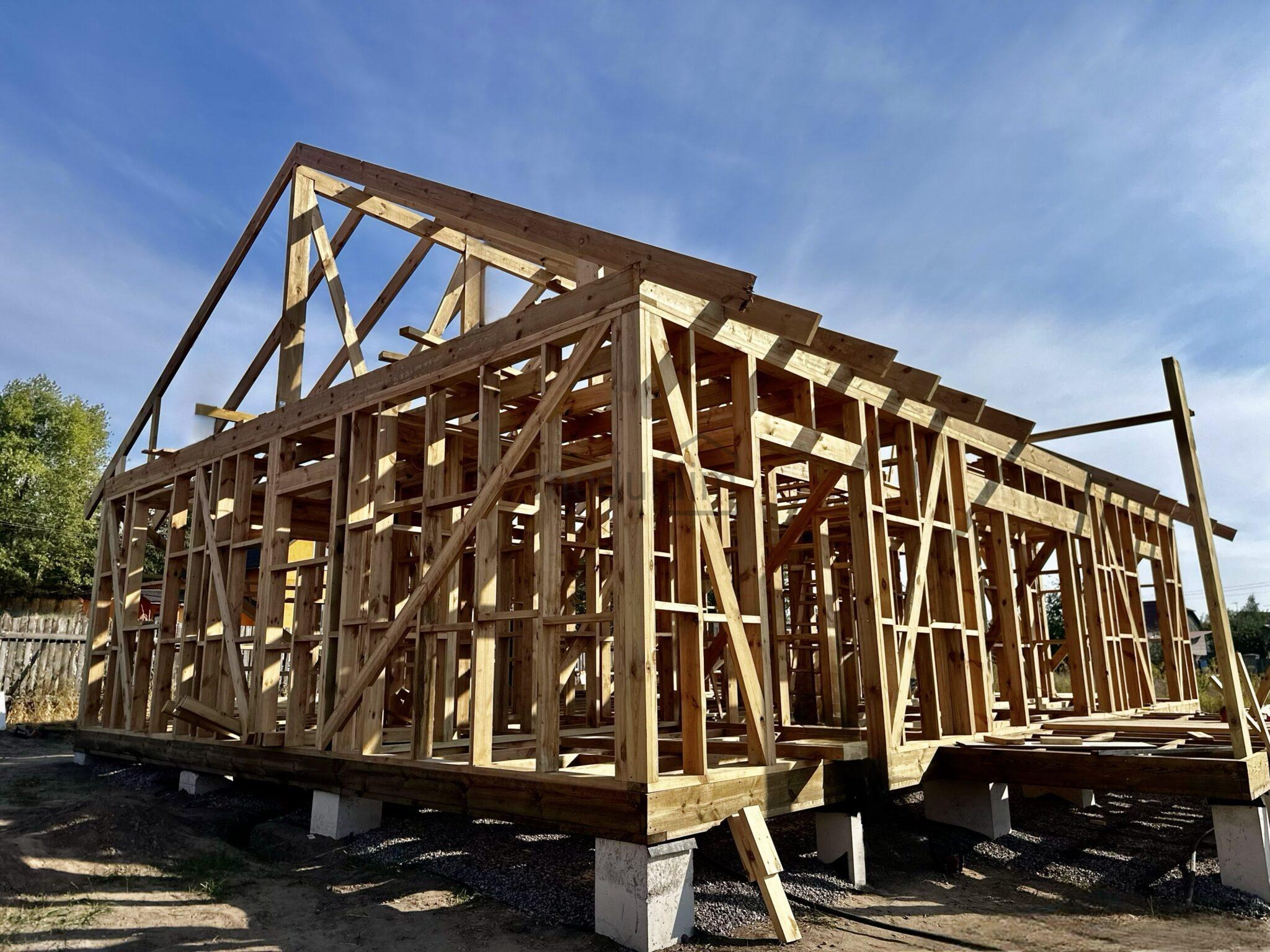 Szkielet drewnianego domu w budowie, widoczny na betonowych fundamentach, konstrukcja dachu w trakcie montażu, błękitne niebo w tle, surowy stan.