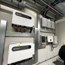 AK POL-ENERGIA SPÓŁKA Z OGRANICZONĄ ODPOWIEDZIALNOŚCIĄ - Dwa białe inwertery Growatt na metalowych szynach, okablowanie na ścianie w szarym pomieszczeniu. Widoczne kable i puszki elektryczne na suficie.