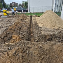 AK POL-ENERGIA SPÓŁKA Z OGRANICZONĄ ODPOWIEDZIALNOŚCIĄ - Wykop pod instalację przy budynku w trakcie prac ziemnych. Widoczny rów, hałda piasku i częściowo mała koparka Volvo. Prace ziemne dla instalacji.