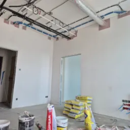 Wnętrze w trakcie remontu: widoczne instalacje pod sufitem, worki z zaprawą, narzędzia i kable. Sufit z instalacjami, ściany przygotowane do wykończenia.