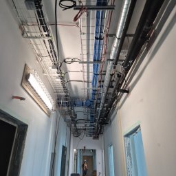 AK POL-ENERGIA SPÓŁKA Z OGRANICZONĄ ODPOWIEDZIALNOŚCIĄ - Korytarz w trakcie prac instalacyjnych: kable w korytkach, rury wentylacyjne pod sufitem. Widoczna osoba w kasku. Oświetlenie sufitowe zamontowane.