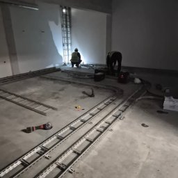 AK POL-ENERGIA SPÓŁKA Z OGRANICZONĄ ODPOWIEDZIALNOŚCIĄ - Montaż tras kablowych w surowym wnętrzu. Dwóch instalatorów pracuje, używając szlifierki kątowej. Widoczne narzędzia i elementy konstrukcyjne tras.