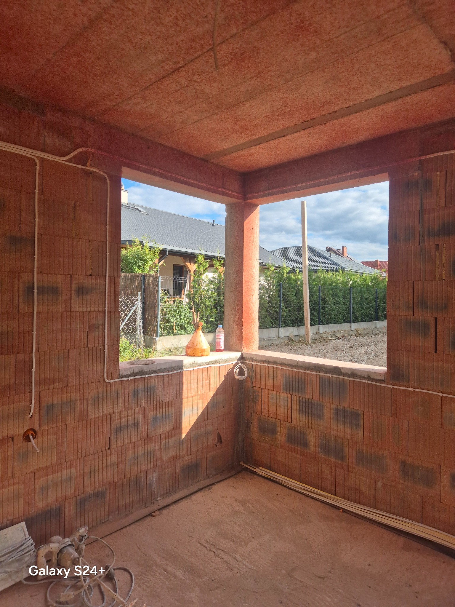 Wnętrze surowego budynku z czerwonej cegły z widoczną instalacją elektryczną w ścianie. Przez okno widać dom i zieleń. Na podłodze leżą narzędzia.