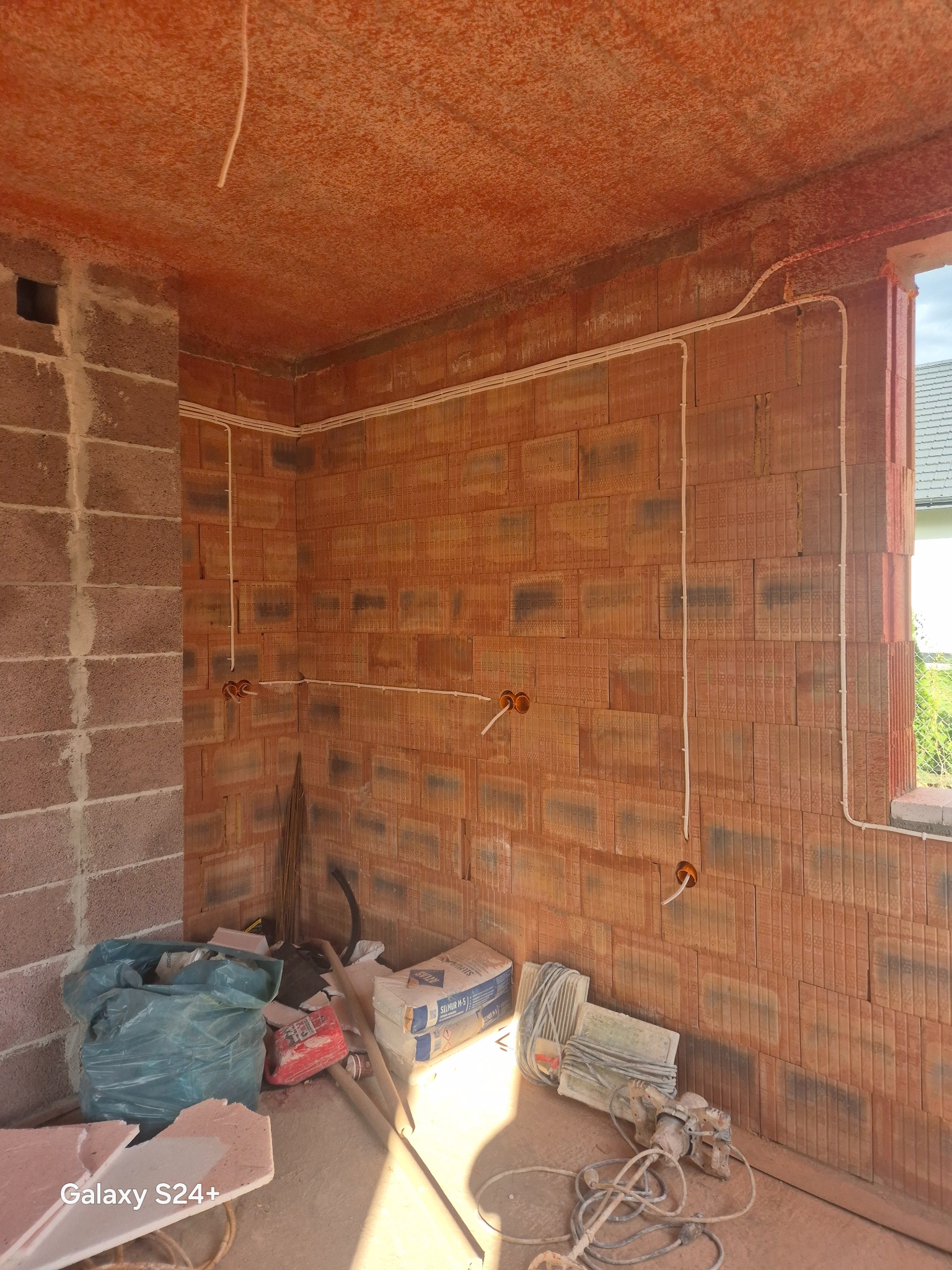 Wnętrze w trakcie prac: ceglane ściany z poprowadzonymi białymi przewodami elektrycznymi, widoczne puszki instalacyjne, gruz i narzędzia na podłodze.