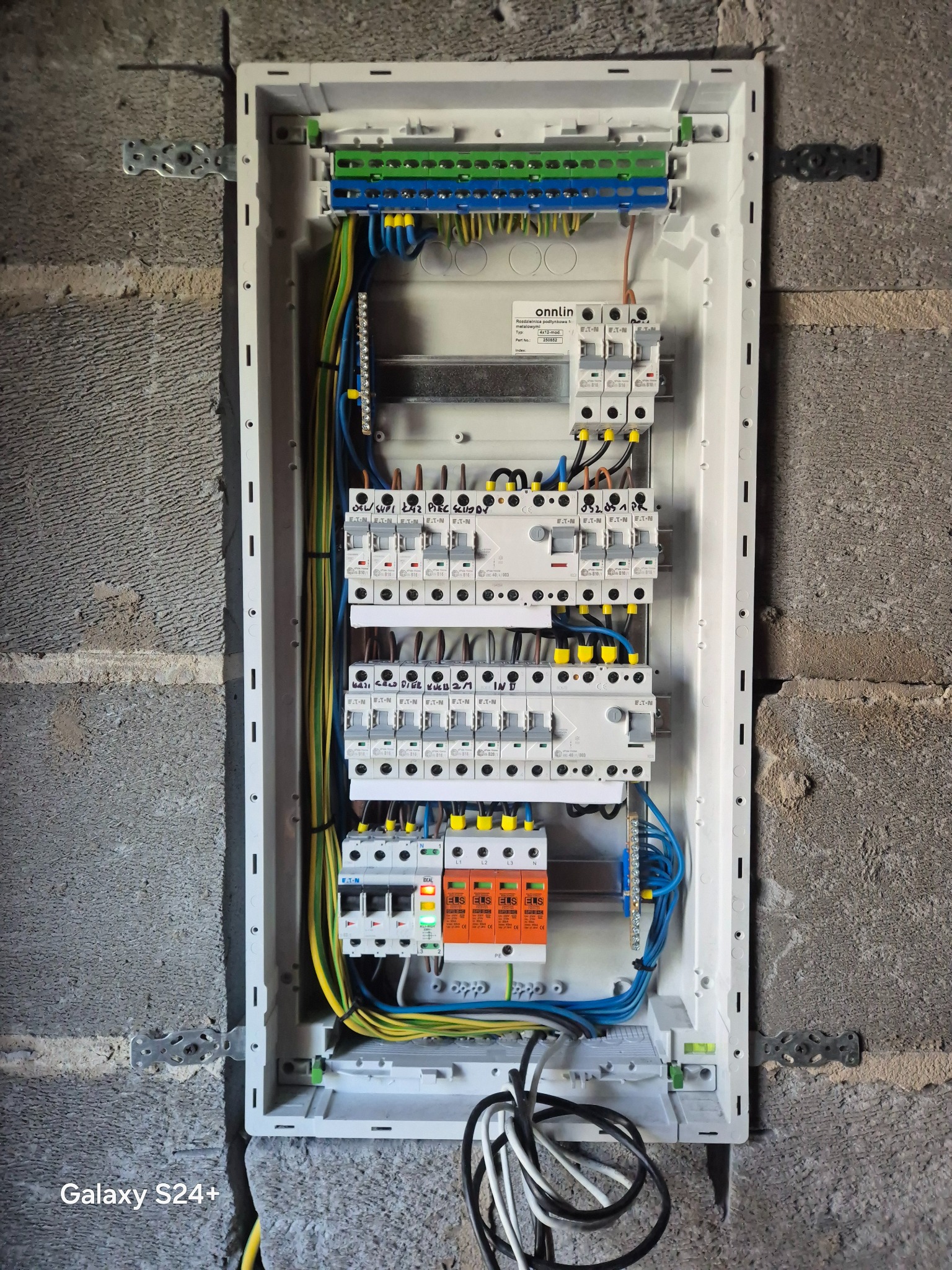 Rozdzielnica elektryczna podtynkowa marki Onnlin, zamontowana w surowej ścianie z betonu komórkowego. Widoczne kable i zabezpieczenia, pomiary i oznaczenia na urządzeniach.