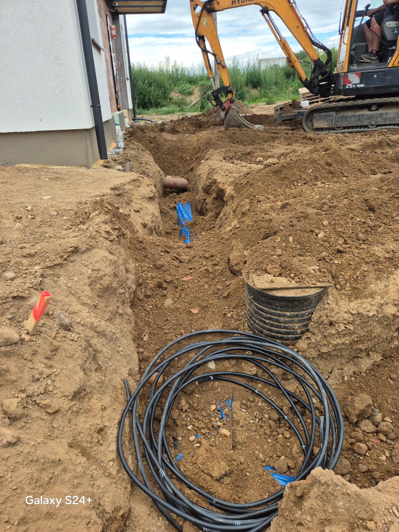 Wykop pod instalacje: rury w ziemi, wykoparka w tle. Widoczne kable i elementy instalacji w wykopie. Prace ziemne w toku.