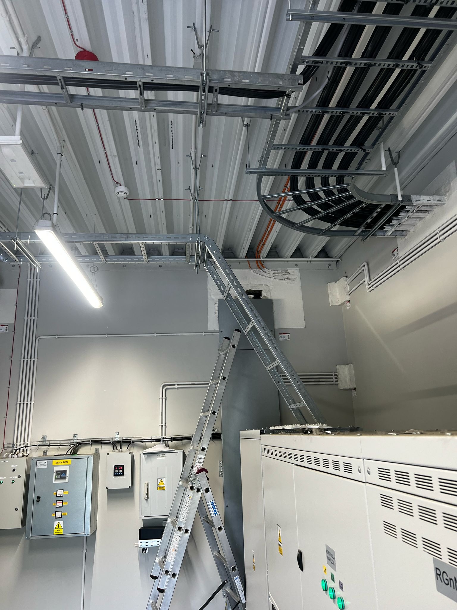 Szczegółowe ujęcie instalacji kablowych pod sufitem, z widoczną drabinką kablową, rurami i szafami elektrycznymi. Aluminiowa drabina oparta o szafę. Oświetlenie LED.