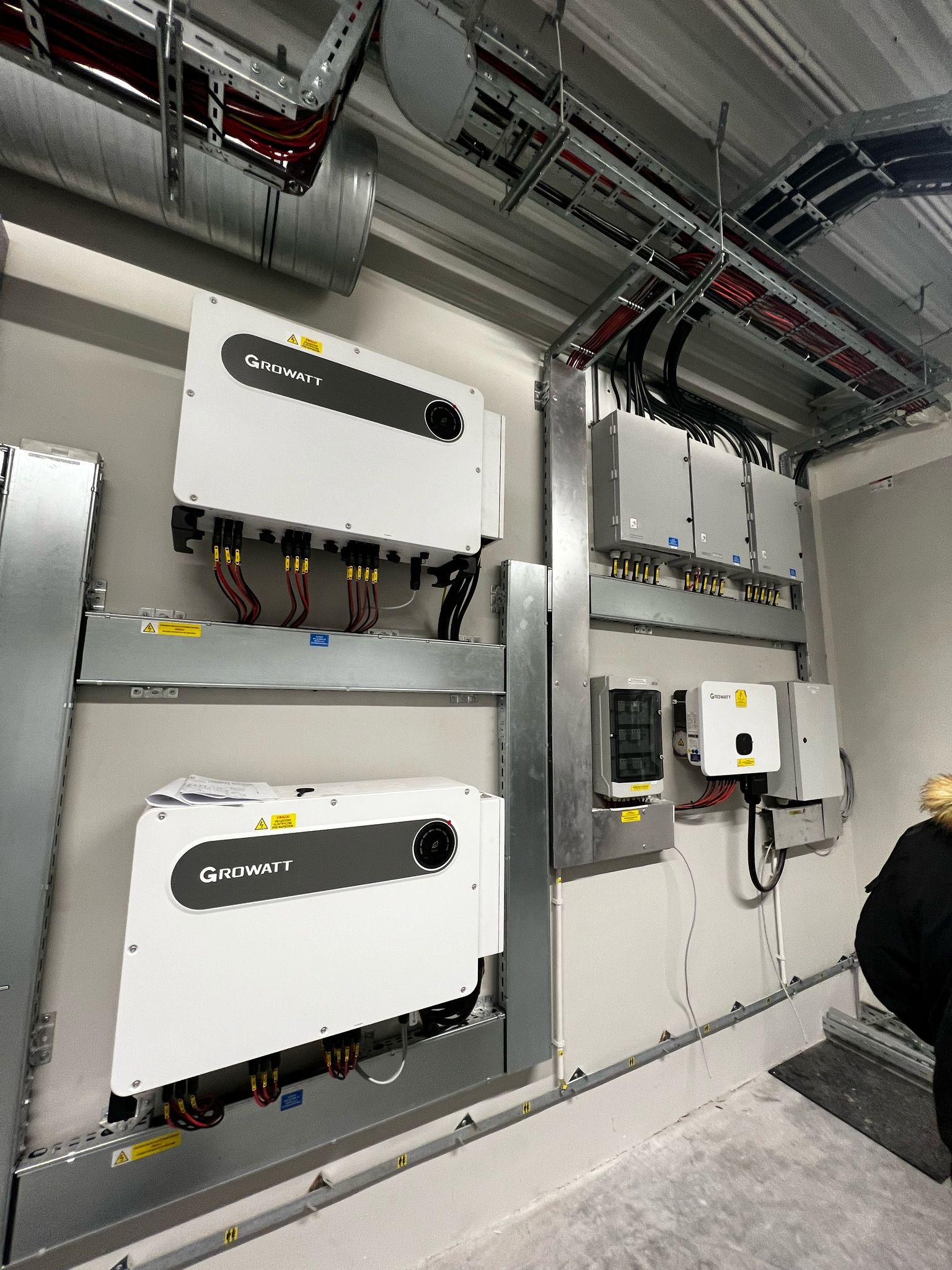 Dwa białe inwertery Growatt na metalowych szynach, okablowanie na ścianie w szarym pomieszczeniu. Widoczne kable i puszki elektryczne na suficie.