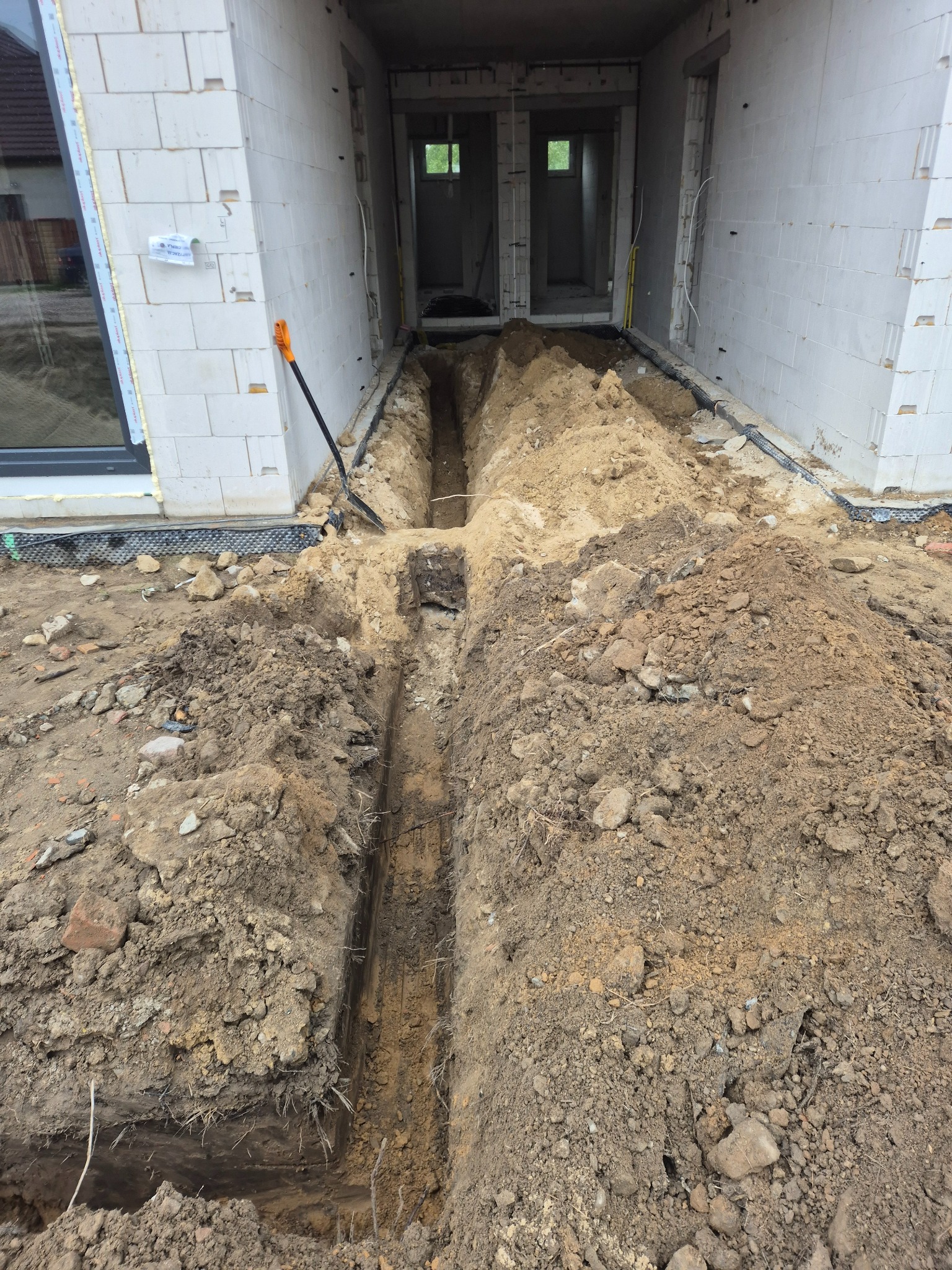 Wykopane rowy pod instalacje elektryczne w nowym budynku z bloczków. Widoczne kable i rury w ścianach. Pomarańczowa łopata oparta o ścianę.