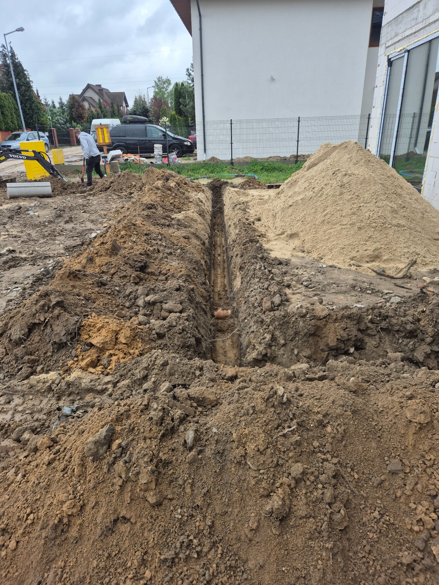 Wykop pod instalację przy budynku w trakcie prac ziemnych. Widoczny rów, hałda piasku i częściowo mała koparka Volvo. Prace ziemne dla instalacji.