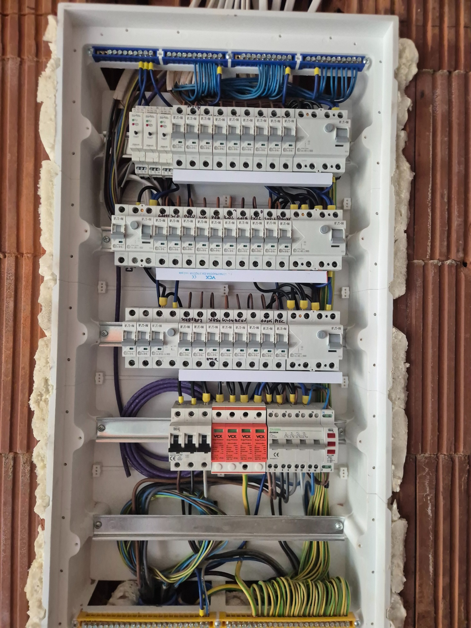 Rozdzielnica elektryczna w białej obudowie, z widocznymi bezpiecznikami, przewodami i listwami zaciskowymi, zamontowana w ścianie z cegły.