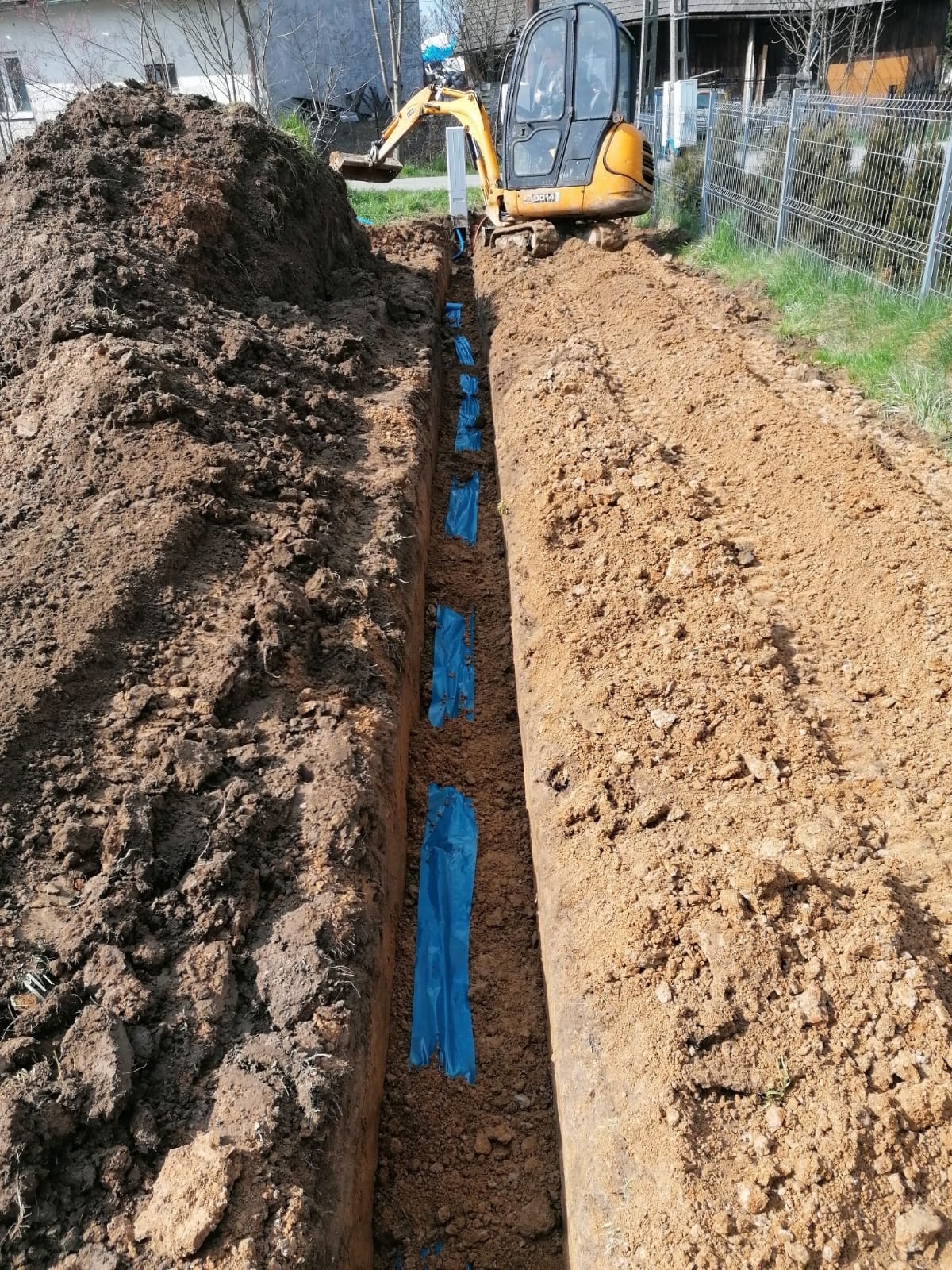 Żółta koparka nad wykopem z niebieską folią zabezpieczającą, przygotowanym pod instalację. Sterta ziemi obok wykopu, siatka ogrodzeniowa w tle.
