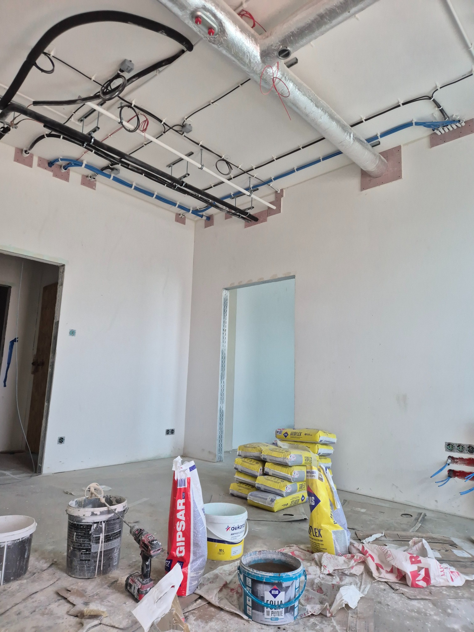 Wnętrze w trakcie remontu: widoczne instalacje pod sufitem, worki z zaprawą, narzędzia i kable. Sufit z instalacjami, ściany przygotowane do wykończenia.