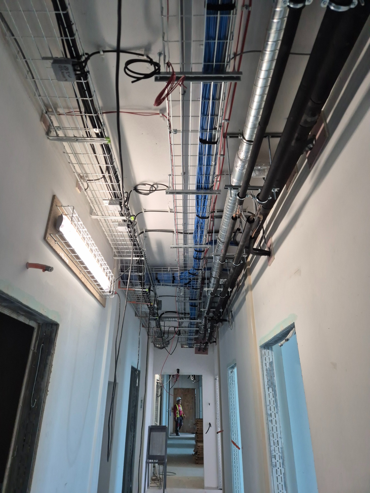 Korytarz w trakcie prac instalacyjnych: kable w korytkach, rury wentylacyjne pod sufitem. Widoczna osoba w kasku. Oświetlenie sufitowe zamontowane.