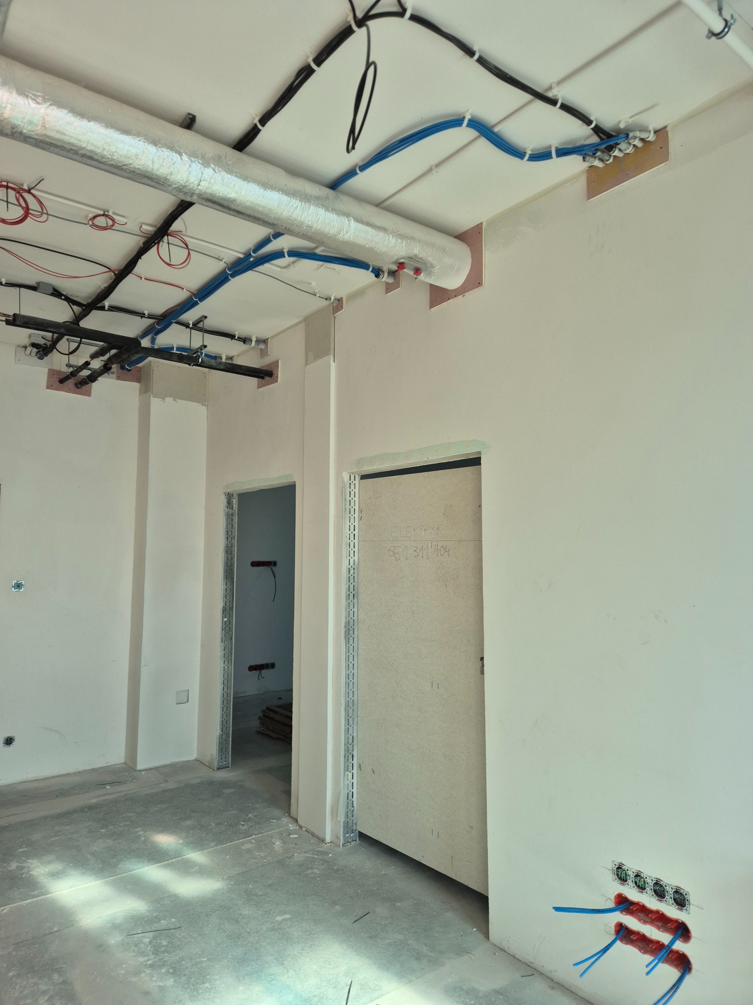 Wnętrze w trakcie prac instalacyjnych: widoczne kable elektryczne w peszlach na suficie, puszki podtynkowe w ścianie, surowy beton i przygotowane otwory drzwiowe.
