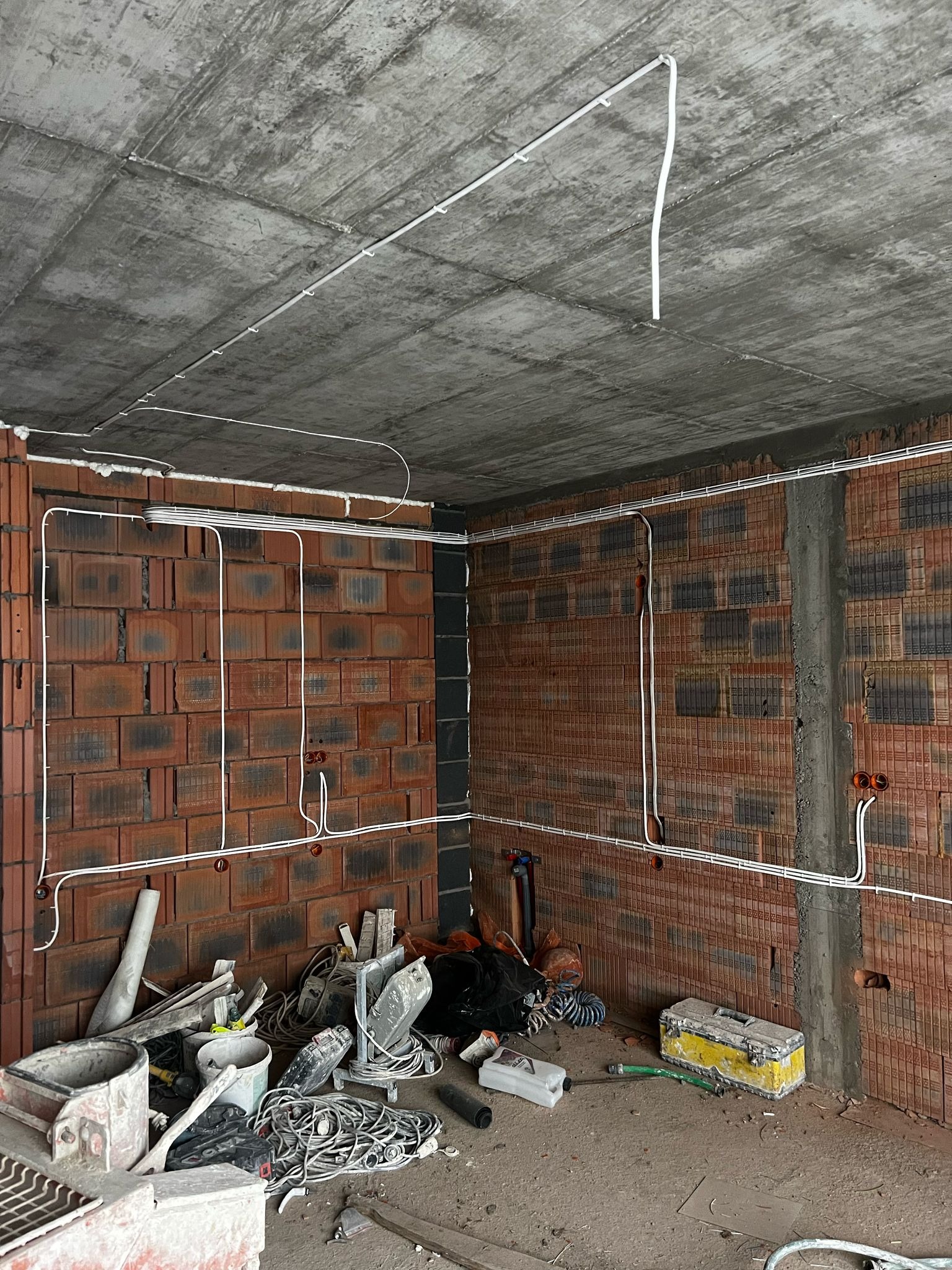 Surowy pokój z ceglanymi ścianami, gdzie widoczna jest instalacja elektryczna w białych rurkach, jeszcze przed wykończeniem. Na podłodze leżą narzędzia.