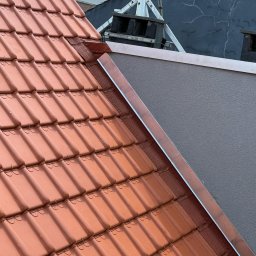 ARTYS - Czerwona dachówka ceramiczna na skośnym dachu, z widoczną metalową obróbką blacharską przy ścianie. W tle drabina dekarska.