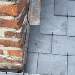 Szczegółowe ujęcie łączenia dachu z kominem, z widocznymi szarymi płytkami dachowymi i obróbką blacharską przy ceglanym kominie. Fragment drewnianej konstrukcji dachu.