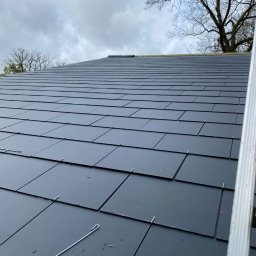 ARTYS - Nowy dach z ciemnej dachówki łuskowej, zabezpieczony metalowymi klamrami. Na tle pochmurne niebo i korona drzewa. Widoczny fragment drabiny.