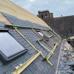 ARTYS - Montaż dachówki na połaci dachu z oknami. W tle widoczna wieża. Fragment rusztowania. Dach pokryty częściowo membraną.