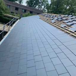 ARTYS - Częściowo pokryty dach z szarych płyt, widoczne drewniane łaty, w tle zieleń drzew i budynek. Prace dekarskie w toku, z bliska.