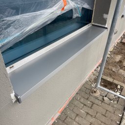 ARTYS - Szary parapet zewnętrzny zamontowany pod oknem, zabezpieczonym folią. Świeżo otynkowana ściana z widocznymi śladami prac budowlanych. Fragment rusztowania.