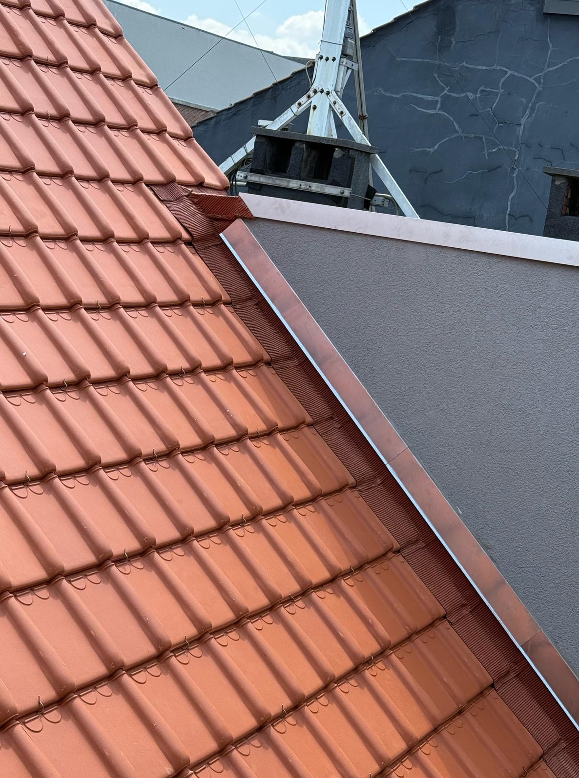 Czerwona dachówka ceramiczna na skośnym dachu, z widoczną metalową obróbką blacharską przy ścianie. W tle drabina dekarska.