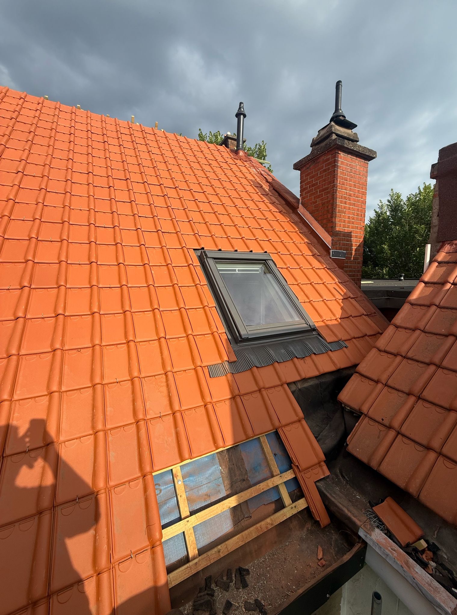 Czerwona dachówka karpiówka z oknem dachowym i kominem. Fragment dachu w trakcie naprawy z widoczną konstrukcją i izolacją. Pochmurne niebo w tle.