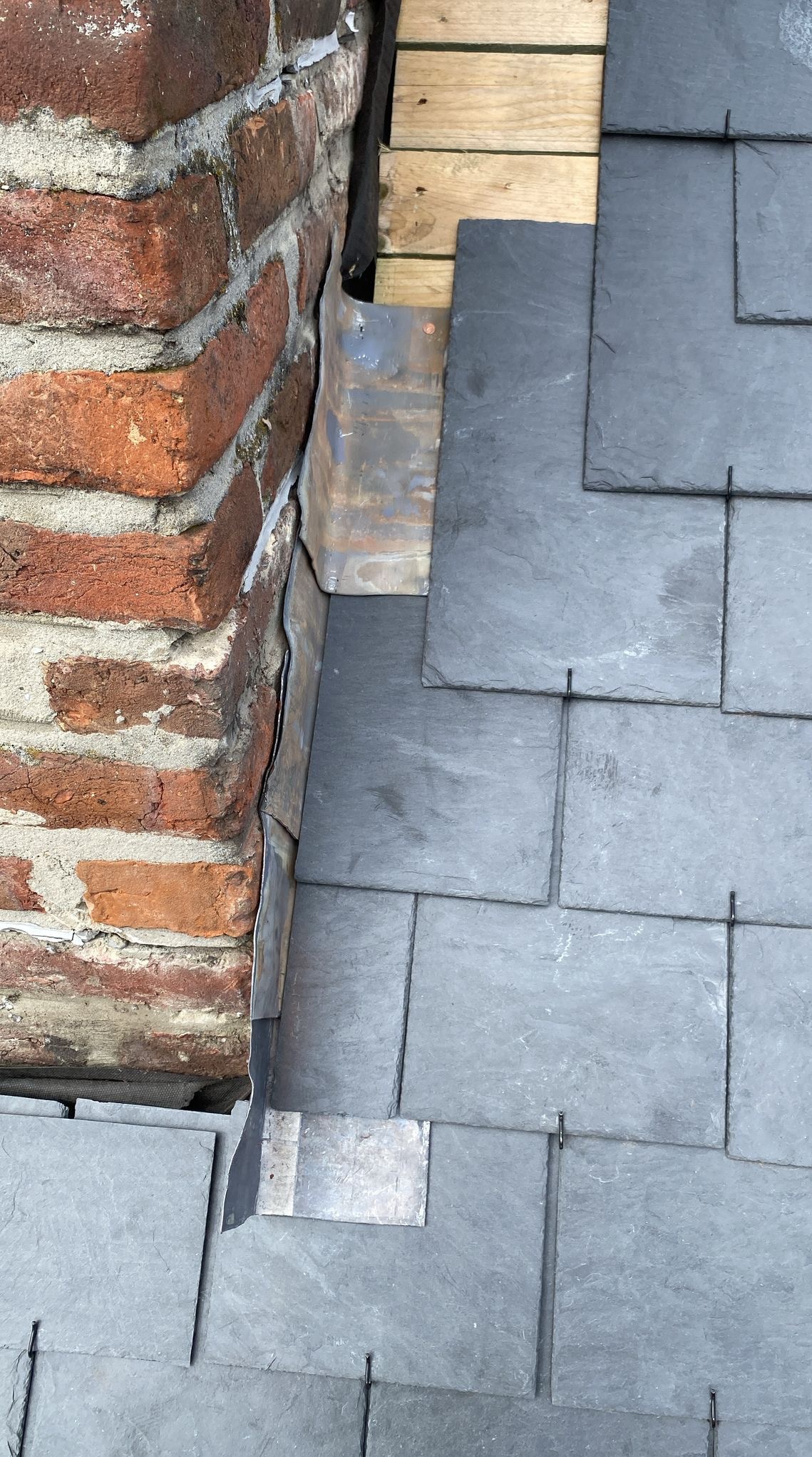 Szczegółowe ujęcie łączenia dachu z kominem, z widocznymi szarymi płytkami dachowymi i obróbką blacharską przy ceglanym kominie. Fragment drewnianej konstrukcji dachu.