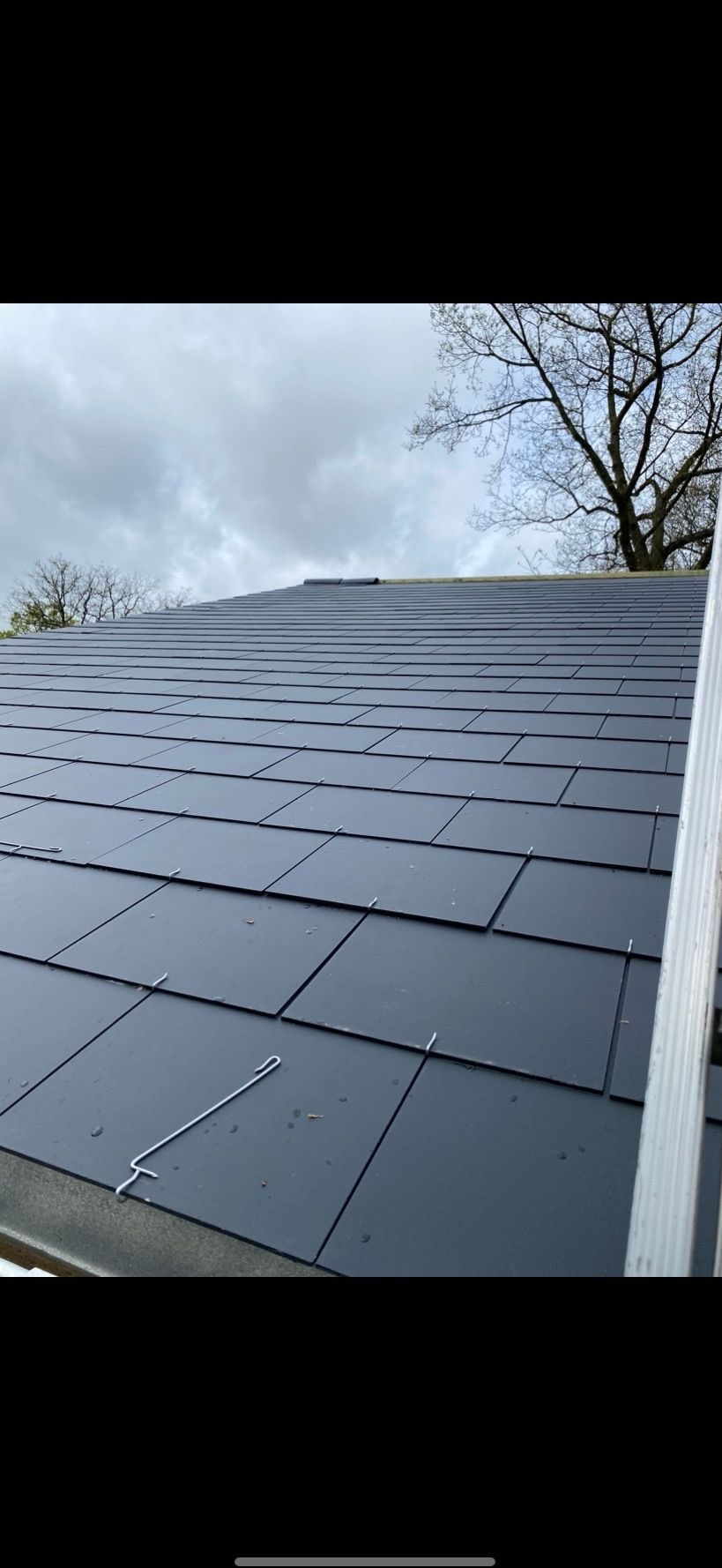 Nowy dach z ciemnej dachówki łuskowej, zabezpieczony metalowymi klamrami. Na tle pochmurne niebo i korona drzewa. Widoczny fragment drabiny.