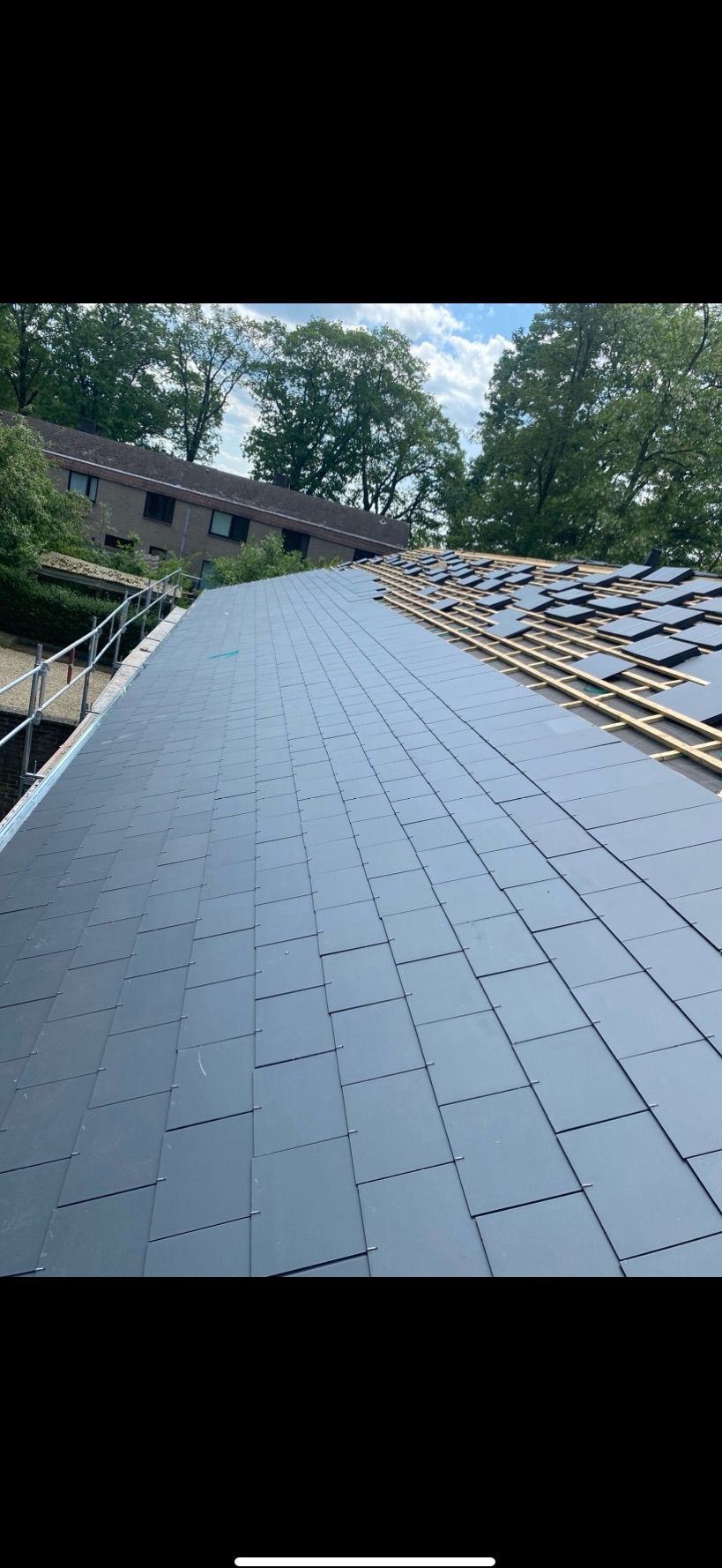 Częściowo pokryty dach z szarych płyt, widoczne drewniane łaty, w tle zieleń drzew i budynek. Prace dekarskie w toku, z bliska.