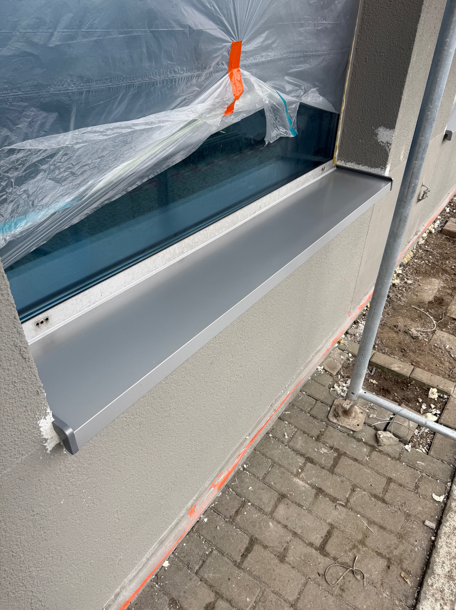 Szary parapet zewnętrzny zamontowany pod oknem, zabezpieczonym folią. Świeżo otynkowana ściana z widocznymi śladami prac budowlanych. Fragment rusztowania.