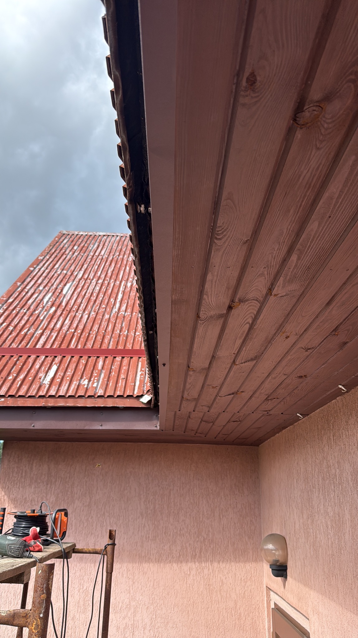Zamontowana, brązowa rynna przy drewnianej podbitce dachowej. W tle blaszany dach i rusztowanie z narzędziami. Ujęcie z dołu, pochmurne niebo.
