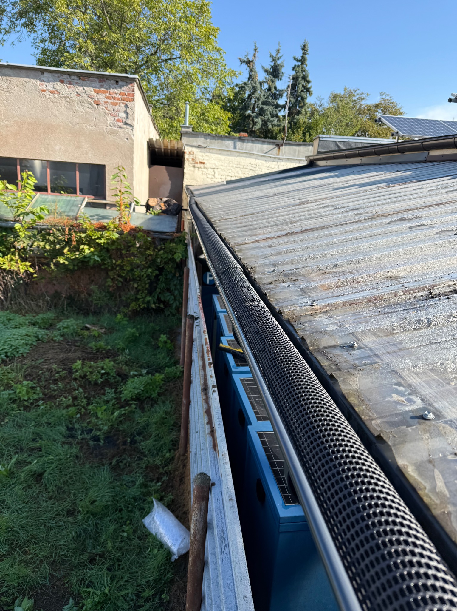 Metalowy dach z zamontowaną czarną osłoną rynny, widok z góry. W tle budynki i zieleń. Rynna zabezpieczona przed liśćmi.