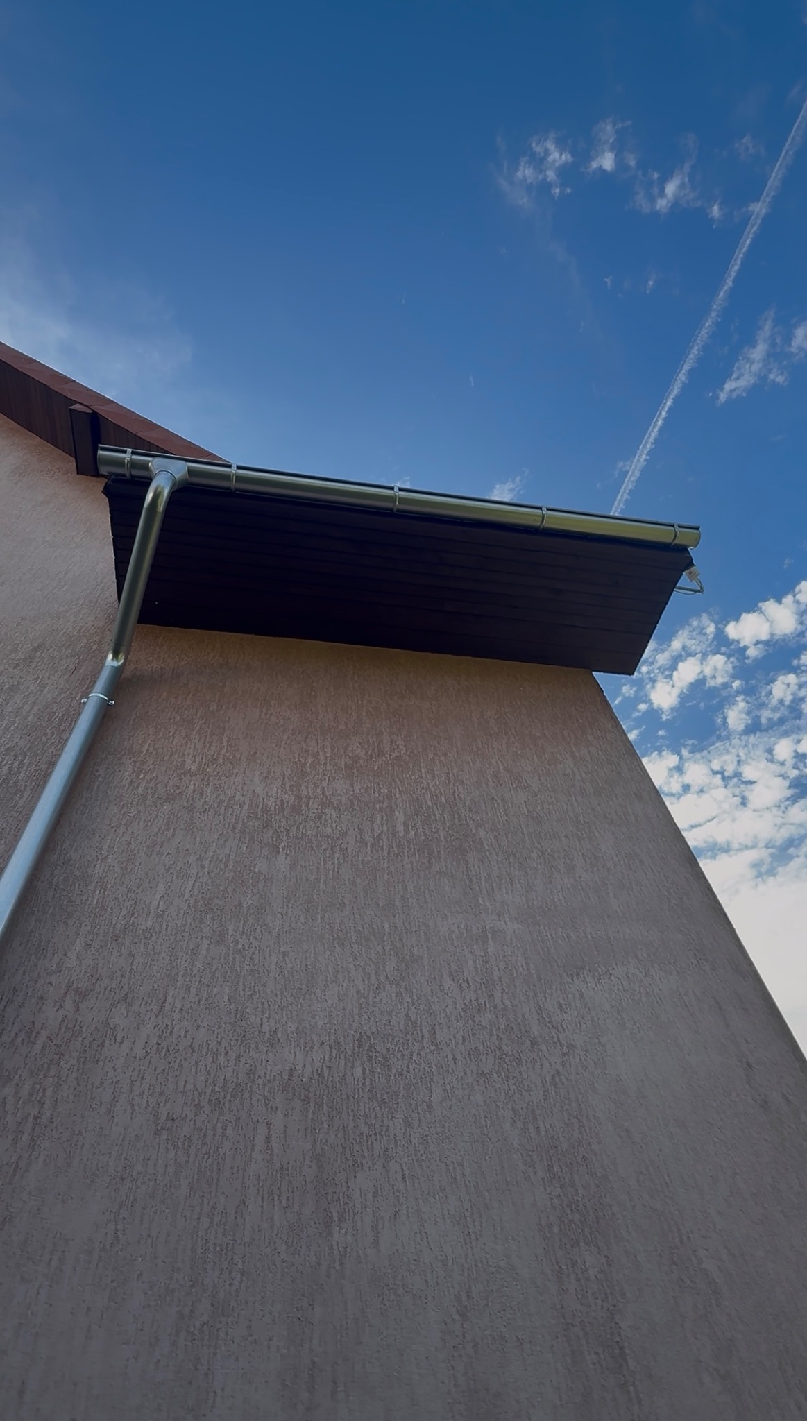 Zamontowana, metalowa rynna pionowa i pozioma na elewacji budynku z brązowym podbitką dachową na tle błękitnego nieba z chmurami.