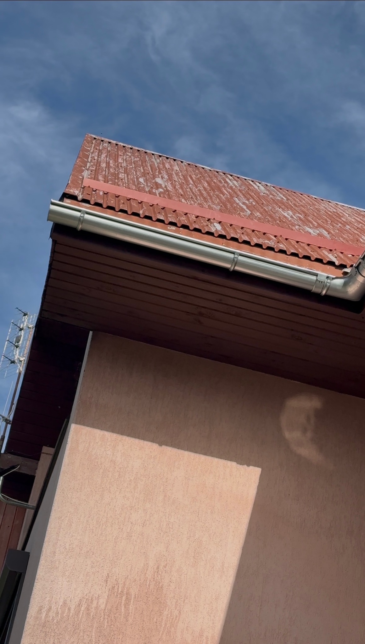 Czerwony dach z blachy falistej, brązowa podbitka i nowa, błyszcząca rynna na tle błękitnego nieba. Ujęcie z dołu, widoczny fragment elewacji budynku.