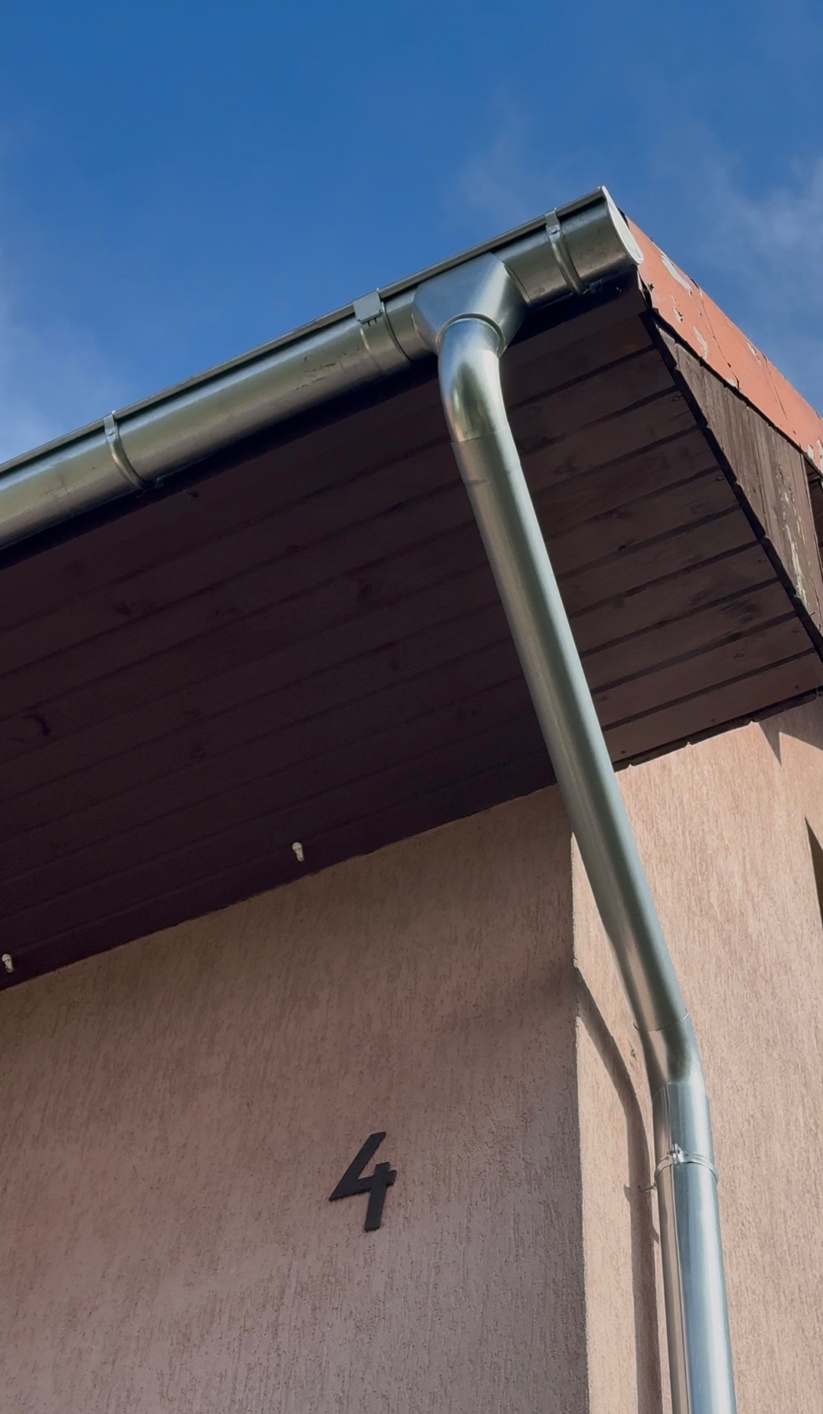 Metalowa rynna spustowa, zamontowana przy brązowym podbitce dachu i beżowej elewacji budynku z numerem 4, na tle błękitnego nieba.