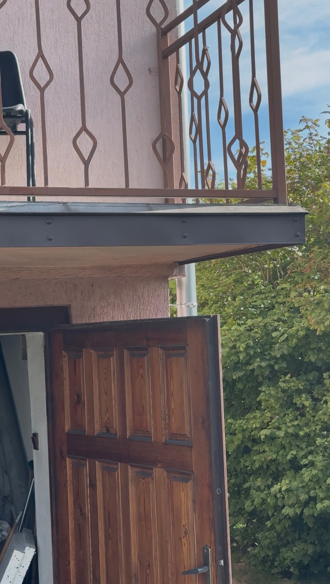 Otwarta drewniana brama balkonowa z metalową balustradą o dekoracyjnym wzorze rombów, na tle zieleni. Widoczne elementy konstrukcyjne balkonu.