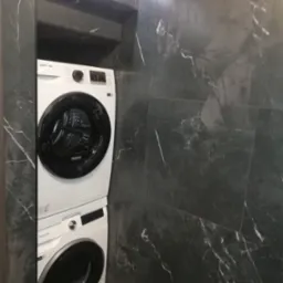 Ustawiona w pionie pralka i suszarka Samsung w łazience wyłożonej ciemnym marmurem z białymi żyłami, oświetlona listwą LED.