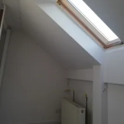 Poddasze z oknem dachowym i grzejnikiem. Białe ściany, drewniana rama okna, jasna podłoga. Widoczne instalacje grzewcze i elektryczne.