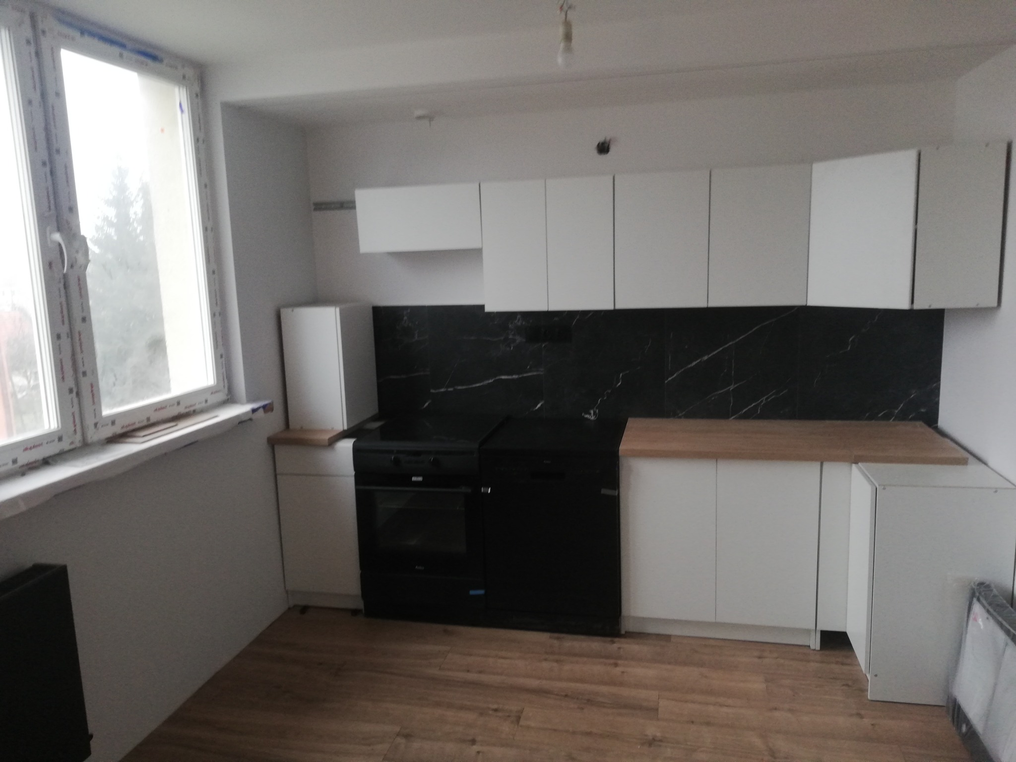 Nowoczesna kuchnia z białymi szafkami i czarnym marmurem na ścianie. Ustawiona jest pod oknem. Na podłodze panele imitujące drewno. Nowe sprzęty AGD w kolorze czarnym.