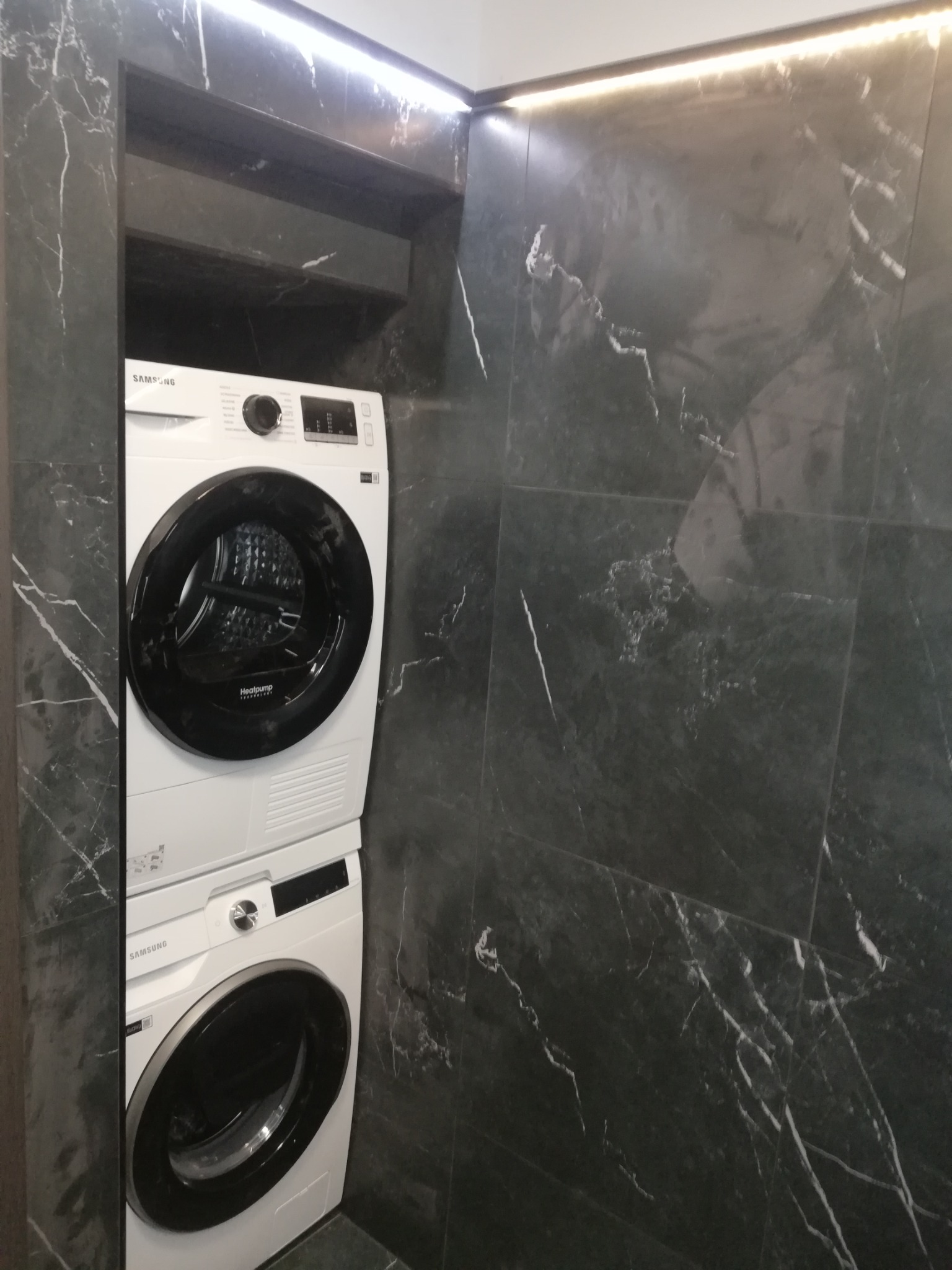 Ustawiona w pionie pralka i suszarka Samsung w łazience wyłożonej ciemnym marmurem z białymi żyłami, oświetlona listwą LED.