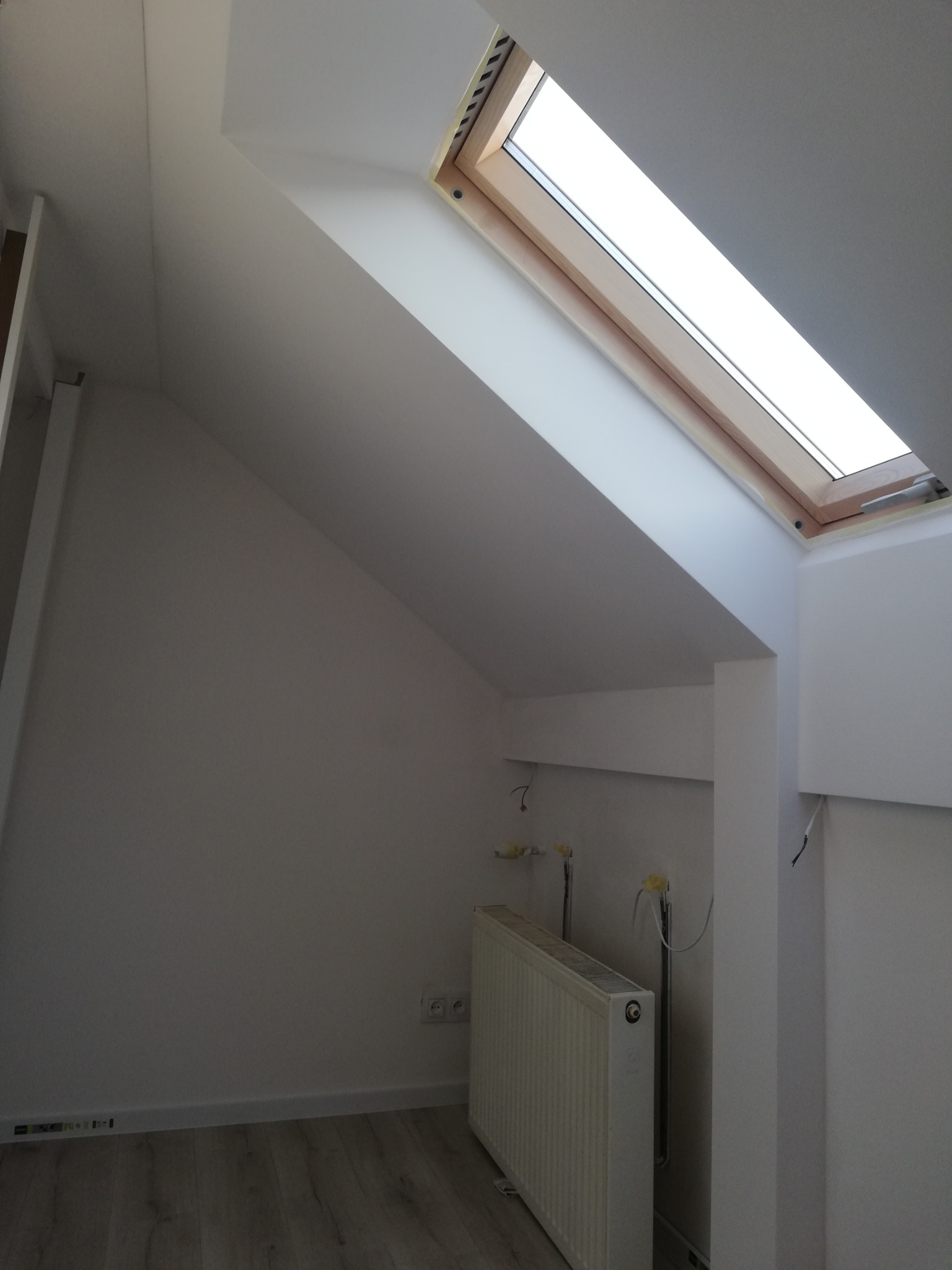 Poddasze z oknem dachowym i grzejnikiem. Białe ściany, drewniana rama okna, jasna podłoga. Widoczne instalacje grzewcze i elektryczne.