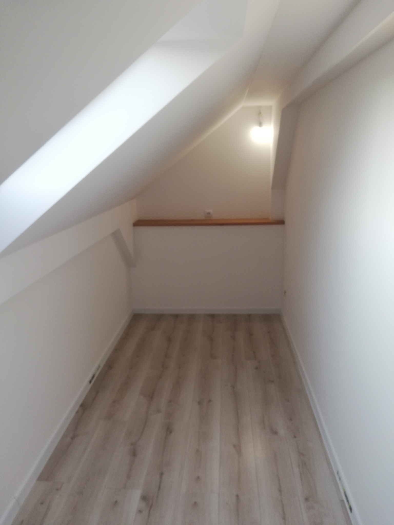 Wykończone wnętrze poddasza ze skosami, świeżo po remoncie. Jasne ściany i podłoga imitująca drewno tworzą przytulną przestrzeń. Widoczna półka pod skosem.