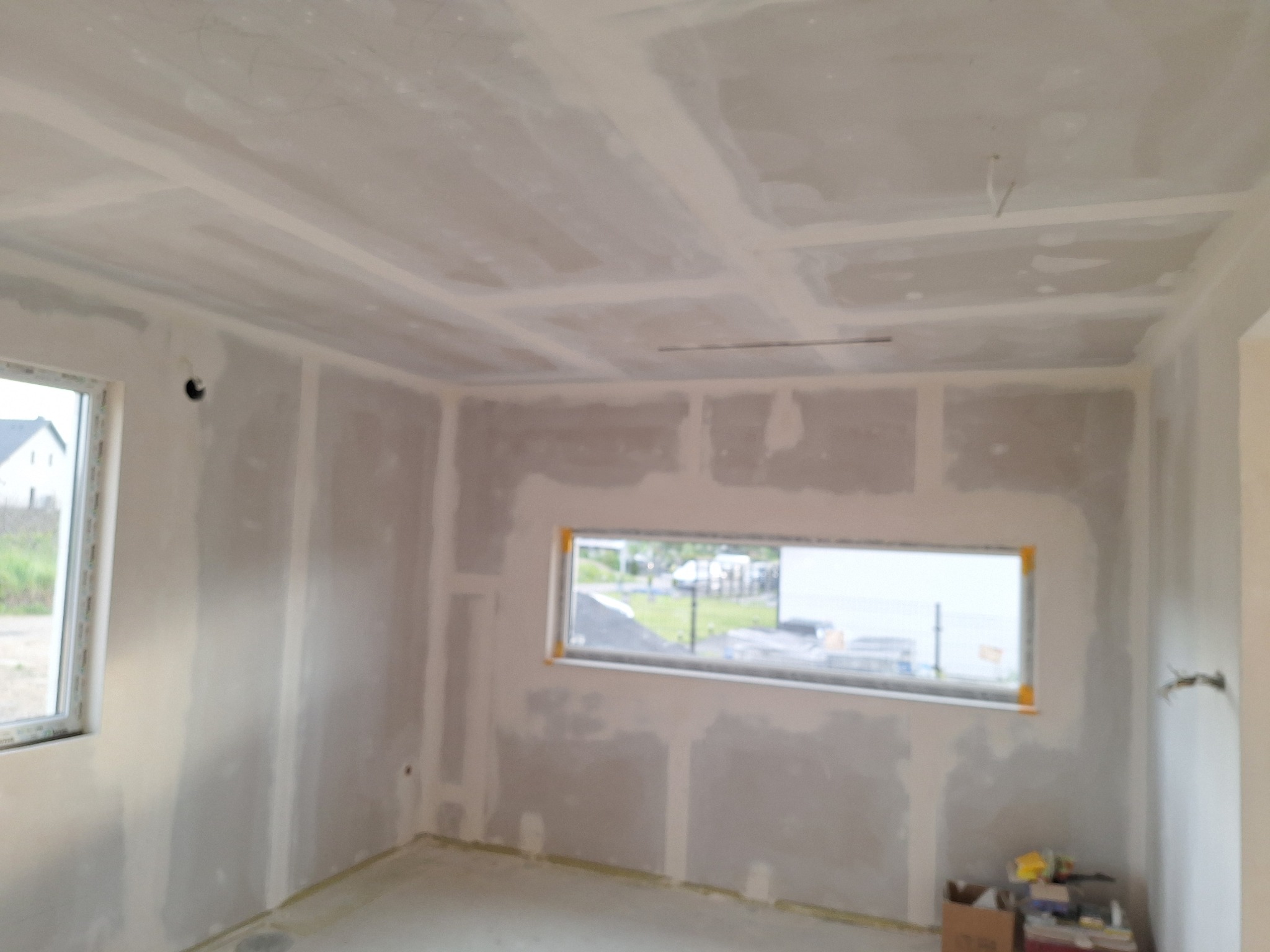 Wnętrze w trakcie prac wykończeniowych: ściany i sufit pokryte płytami gipsowo-kartonowymi, widoczne okna, elementy instalacji elektrycznej. W tle fragment krajobrazu.