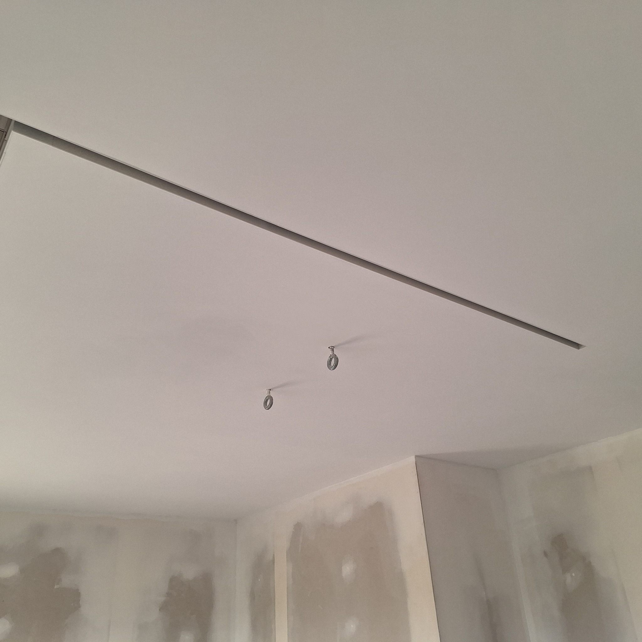 Montaż sufitu podwieszanego z oświetleniem liniowym. Widoczne szyny i punkty mocowania lamp. Ściany przygotowane do malowania.