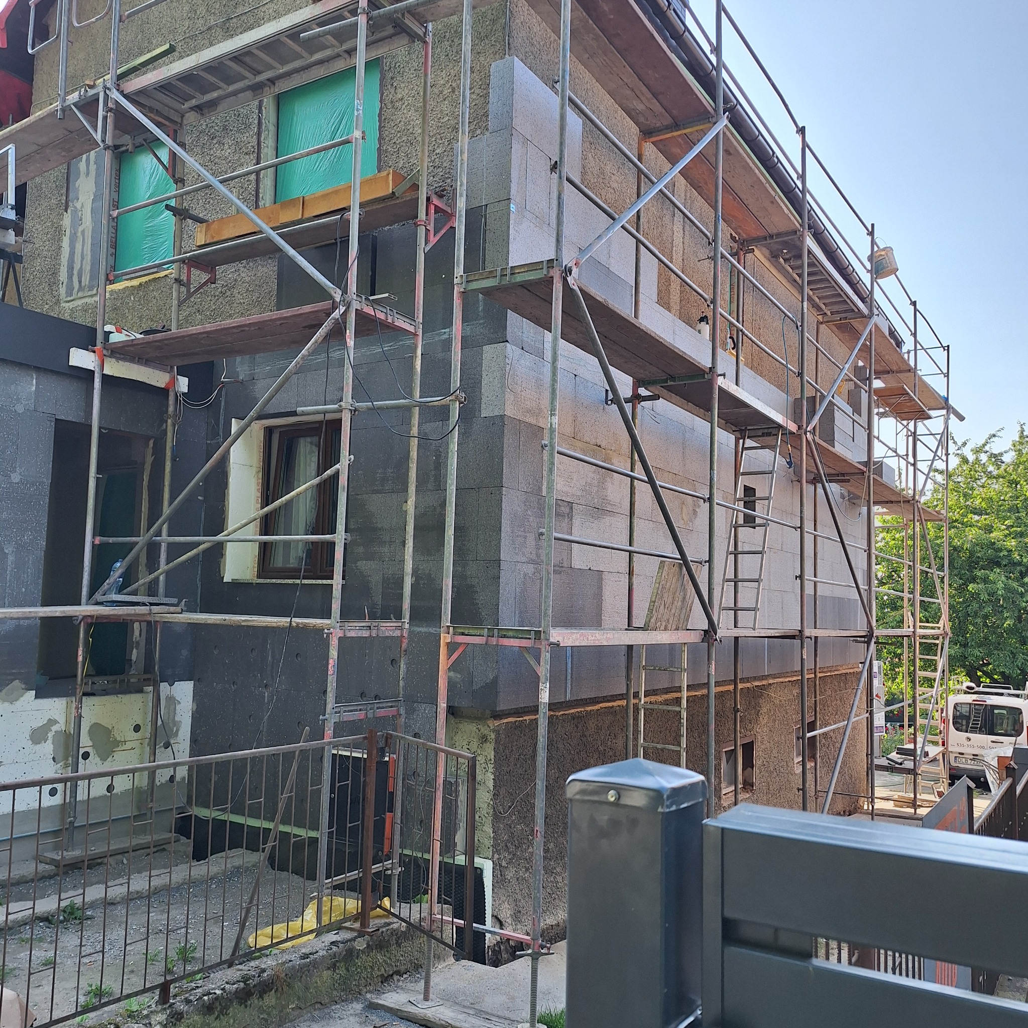 Ocieplanie ścian zewnętrznych domu styropianem grafitowym, widoczne rusztowania i okna zabezpieczone folią. Prace w toku, ściany częściowo ocieplone.